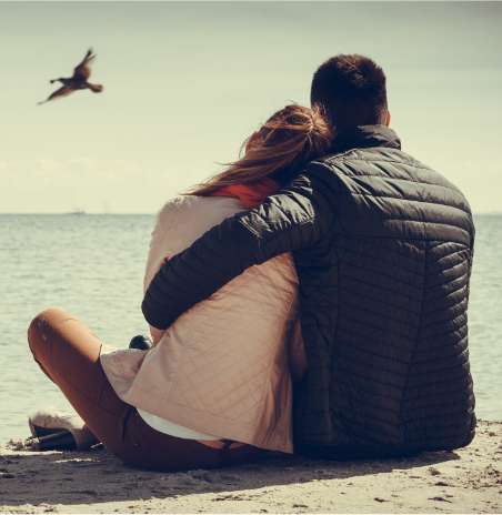 Para w kurtkach przytulona siedzi na plaży.